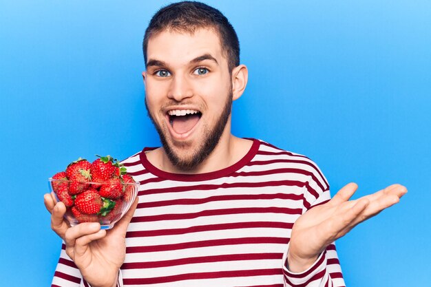 Junger gutaussehender Mann, der Schüssel mit Erdbeeren hält und mit glücklichem Lächeln und Siegerausdruck mit erhobener Hand Leistung feiert