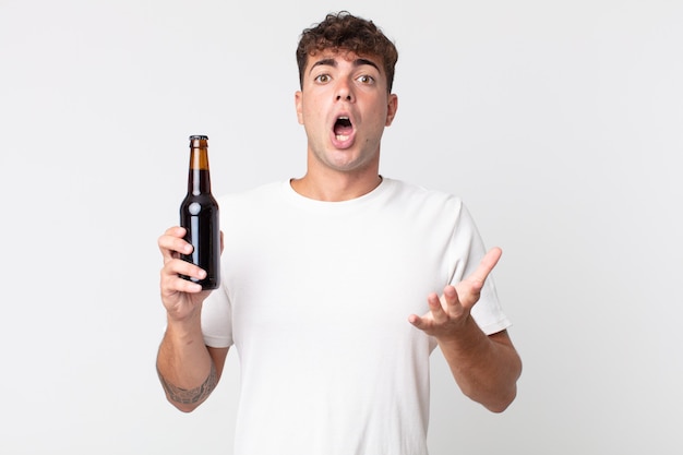 Junger gutaussehender Mann, der sich extrem schockiert und überrascht fühlt und eine Bierflasche hält