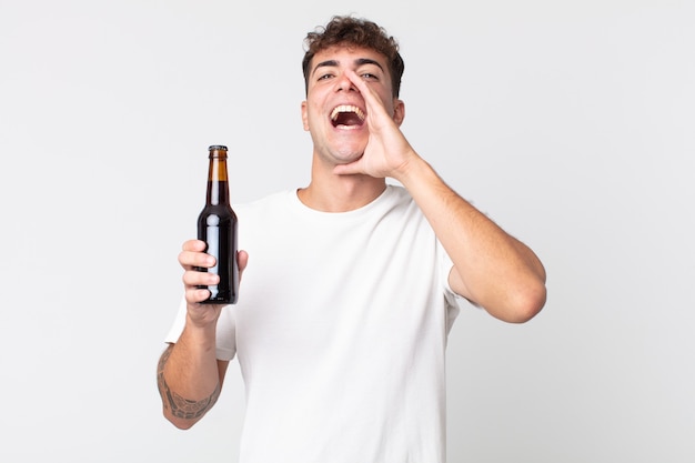 Junger gutaussehender Mann, der sich glücklich fühlt, mit den Händen neben dem Mund einen großen Schrei ausspricht und eine Bierflasche hält