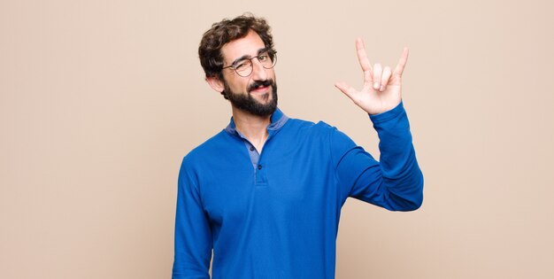 Junger gutaussehender Mann, der sich glücklich, lustig, selbstbewusst, positiv und rebellisch fühlt und Stein- oder Schwermetallzeichen mit Hand gegen flache Wand macht