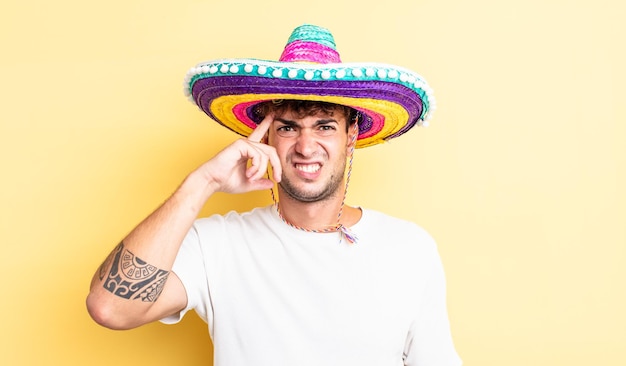 Junger gutaussehender Mann, der sich verwirrt und verwirrt fühlt und zeigt, dass Sie verrückt sind. mexikanisches Hutkonzept