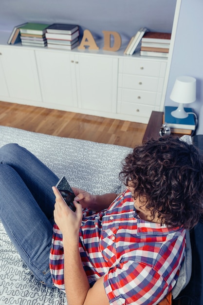 Junger gutaussehender Mann, der Smartphone über einem Bett sitzt. Freizeit zu Hause Konzept.