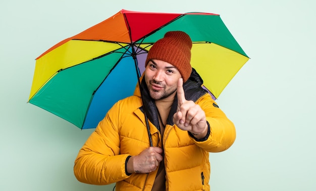 Junger gutaussehender Mann, der stolz und selbstbewusst lächelt und das Regen- und Regenschirmkonzept Nummer eins macht