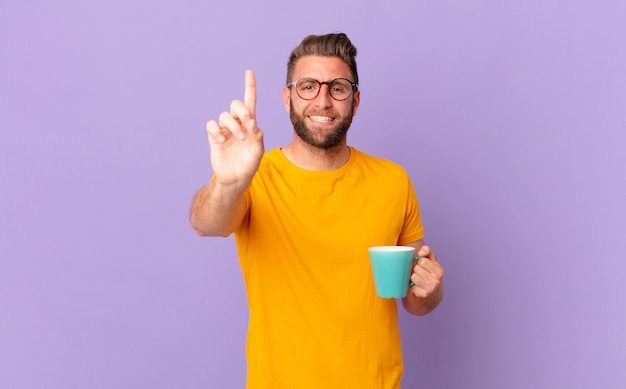 Junger gutaussehender Mann, der stolz und selbstbewusst lächelt und die Nummer eins macht. und hält eine Kaffeetasse