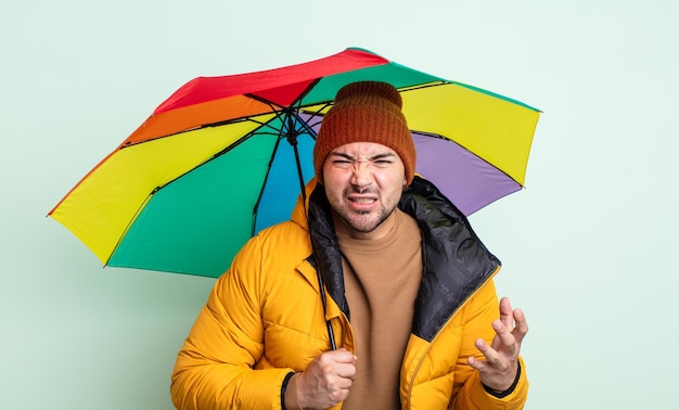 Junger gutaussehender Mann, der wütend, verärgert und frustriert aussieht. Regen- und Regenschirmkonzept