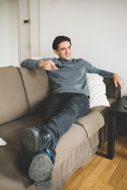 Junger gutaussehender Mann drinnen zu Hause sitzende Couch beim Fernsehen mit Fernbedienung