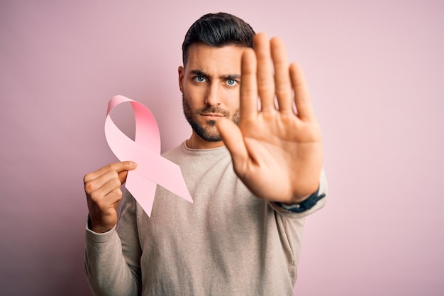 Junger gutaussehender Mann hält ein rosafarbenes Krebsband als Symbol der Unterstützung vor isoliertem Hintergrund und macht mit offener Hand ein Stoppschild mit ernsthafter und selbstbewusster Gesichtsausdruck-Verteidigungsgeste