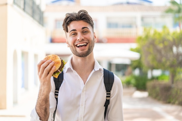 Junger, gutaussehender Mann hält im Freien mit Überraschung und schockiertem Gesichtsausdruck einen Burger in der Hand