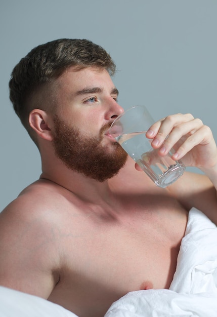 Junger gutaussehender Mann im Bett im Schlafzimmer zu Hause, der morgens unter einer weißen Decke liegt und reines Süßwasser aus Glas trinkt