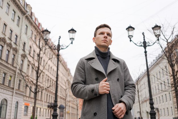 Junger gutaussehender Mann im grauen Mantel geht bei grauem Wetter auf der Straße in der Stadt spazieren