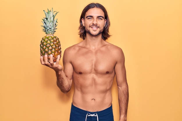 Junger gutaussehender Mann in Badebekleidung, der eine Ananas in der Hand hält, sieht positiv und glücklich aus, steht und lächelt mit einem selbstbewussten Lächeln, das Zähne zeigt
