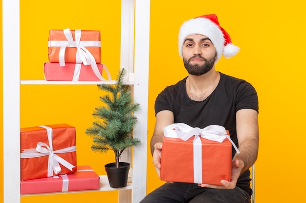 Junger gutaussehender Mann in einem Weihnachtsmann-Hut hält Glückwunschgeschenke neben einem Weihnachtsbaum. Konzept der Feiertage von Weihnachten und Neujahr.