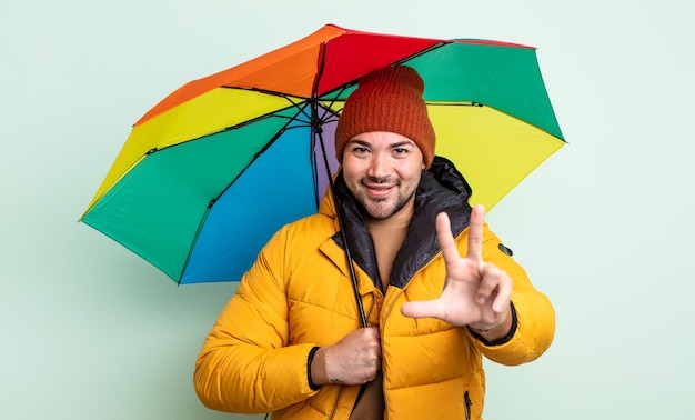 Junger gutaussehender Mann lächelt und sieht freundlich aus und zeigt das Regen- und Regenschirmkonzept Nummer drei