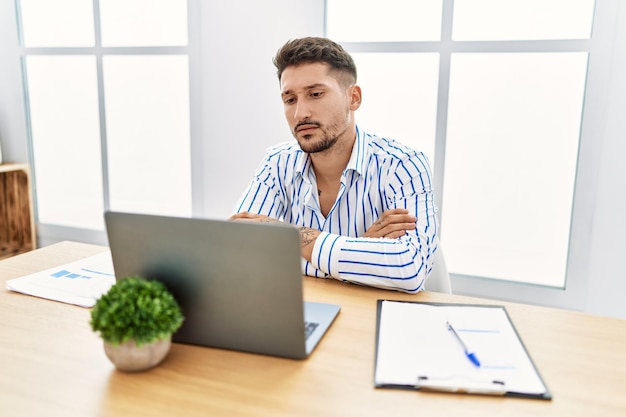 Junger gutaussehender Mann mit Bart, der im Büro mit einem Computer-Laptop arbeitet, skeptisch und nervöser, missbilligender Gesichtsausdruck mit verschränkten Armen