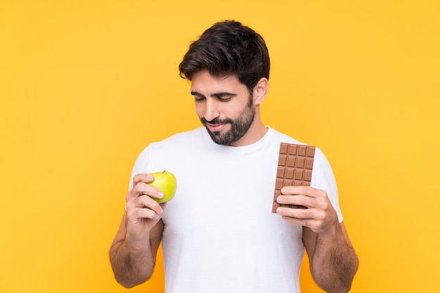 Junger gutaussehender Mann mit Bart über der lokalisierten gelben Wand, die eine Schokoladentablette in einer Hand und einen Apfel in der anderen nimmt