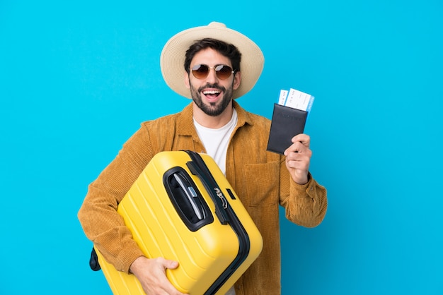Junger gutaussehender Mann mit Bart über lokalisierter blauer Wand in den Ferien mit Koffer und Pass