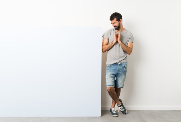 Junger gutaussehender Mann mit dem Bart, der ein großes blaues leeres Plakat entwirft etwas hält