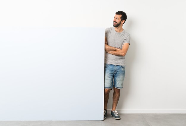 Junger gutaussehender Mann mit dem Bart, der ein großes blaues leeres Plakat glücklich und das Lächeln hält