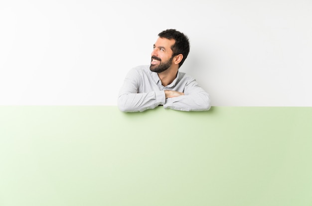 Junger gutaussehender Mann mit dem Bart, der ein großes grünes leeres Plakat glücklich und das Lächeln hält