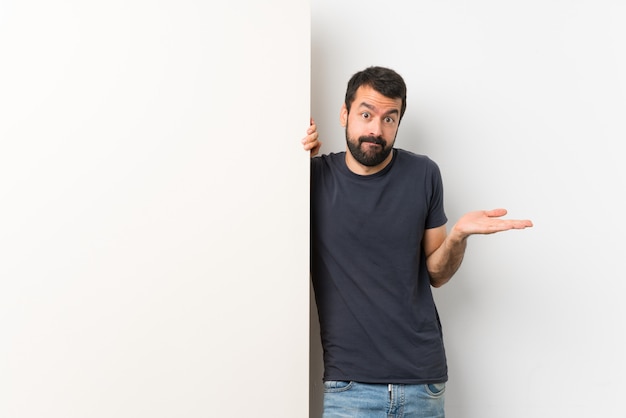 Junger gutaussehender Mann mit dem Bart, der ein großes leeres Plakat hat Zweifel beim Anheben von Händen hält