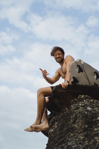 Junger gutaussehender Mann mit einem Surfbrett auf einem Felsen in der Nähe des Ozeans.