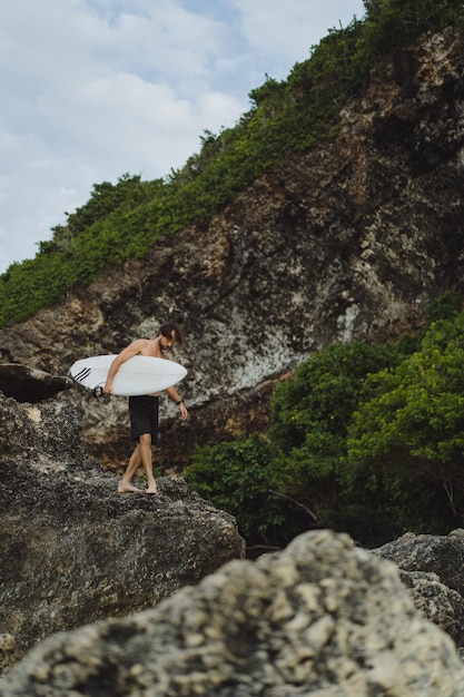 Junger gutaussehender Mann mit einem Surfbrett auf einem Felsen in der Nähe des Ozeans.