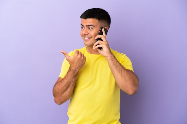 Junger gutaussehender Mann mit Handy über isoliertem lila Hintergrund, der auf die Seite zeigt, um ein Produkt zu präsentieren