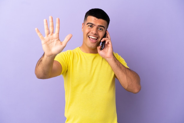 Junger gutaussehender Mann mit Handy über isoliertem lila Hintergrund, der fünf mit den Fingern zählt