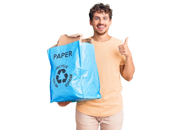 Junger gutaussehender Mann mit lockigem Haar, der Recycling-Papierkorb mit Papier und Pappe hält, der glücklich und positiv mit dem Daumen nach oben lächelt und ausgezeichnetes und Genehmigungszeichen tut