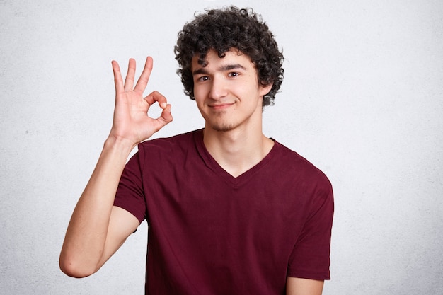 Junger gutaussehender Mann mit lockigem Haar, macht okay Zeichen, zeigt seine Zustimmung, Gesten mit der Hand, isoliert über Weiß