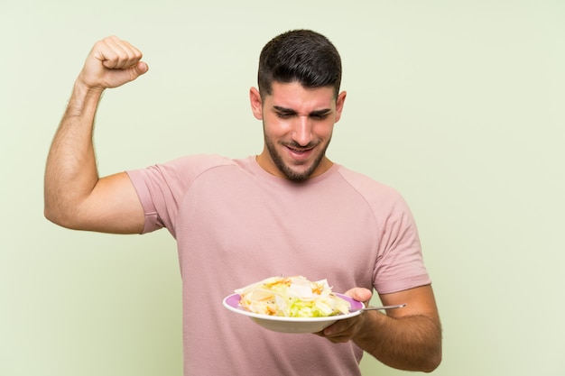 Junger gutaussehender Mann mit Salat über der lokalisierten grünen Wand, die einen Sieg feiert