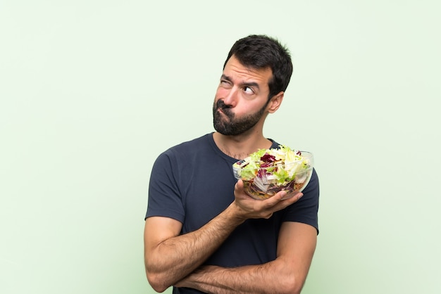 Junger gutaussehender Mann mit Salat über grüner Wand mit verwirren Gesichtsausdruck