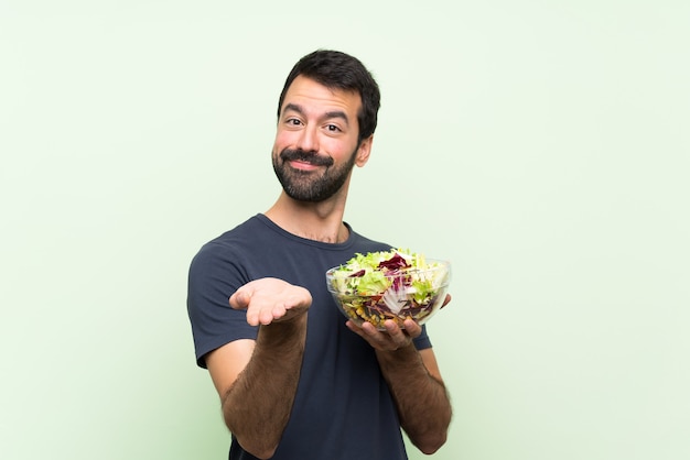 Junger gutaussehender Mann mit Salat über lokalisierter grüner Wand, die copyspace eingebildet auf der Palme hält, um eine Anzeige einzufügen