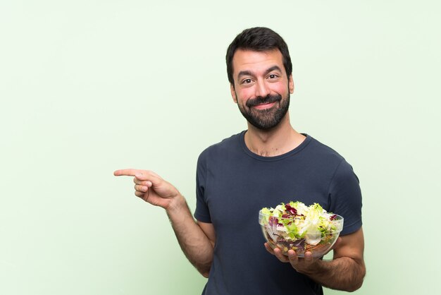 Junger gutaussehender Mann mit Salat über lokalisierter grüner Wand Finger auf die Seite zeigend