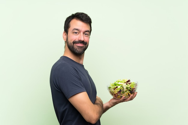 Junger gutaussehender Mann mit Salat über lokalisierter grüner Wand mit den Armen gekreuzt und vorwärts schauend