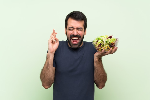 Junger gutaussehender Mann mit Salat über lokalisierter grüner Wand mit der Fingerüberfahrt