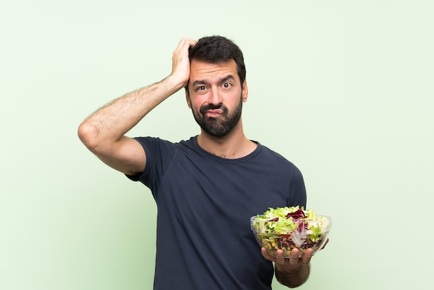Junger gutaussehender Mann mit Salat über lokalisierter grüner Wand mit einem Ausdruck der Frustration und des Nichtverständnisses