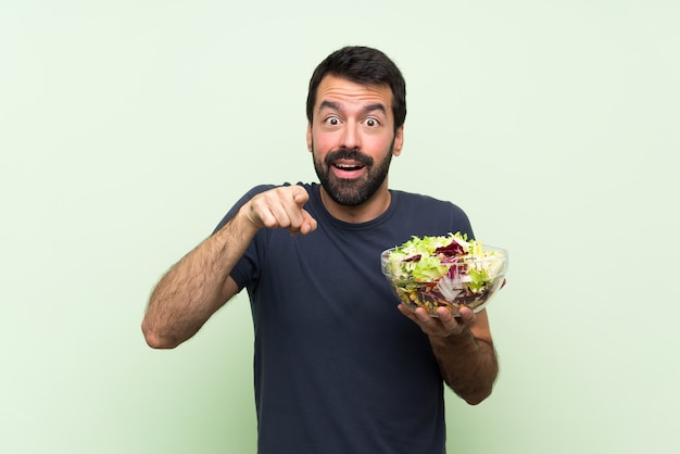 Junger gutaussehender Mann mit Salat über lokalisierter grüner Wand überrascht und Front zeigend