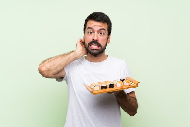 Junger gutaussehender Mann mit Sushi über der lokalisierten grünen Wand, die Zweifel hat