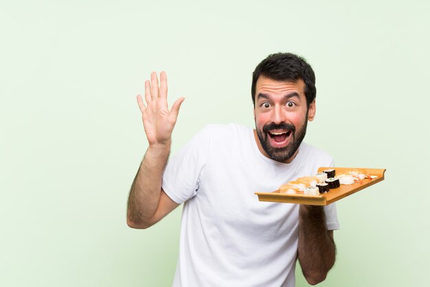 Junger gutaussehender Mann mit Sushi über der lokalisierten grünen Wand nervös und erschrocken