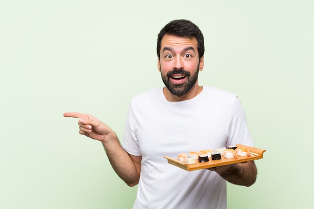 Junger gutaussehender Mann mit Sushi über lokalisierter grüner Wand überraschte und Finger auf die Seite zeigend