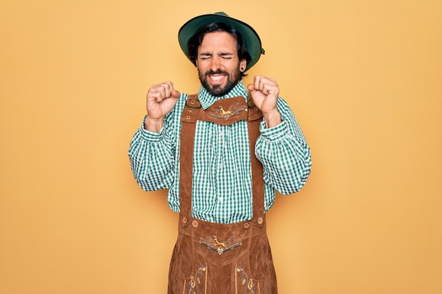 Junger gutaussehender Mann mit traditionellem deutschem Oktoberfest-Custome für Deutschland-Festival, aufgeregt über den Erfolg mit erhobenen Armen und geschlossenen Augen, um den Sieg zu feiern, lächelndes Siegerkonzept