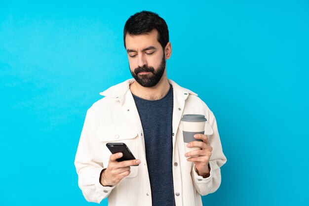 Junger gutaussehender Mann mit weißer Cordjacke über isolierter blauer Wand, die Kaffee hält, um und ein Handy wegzunehmen