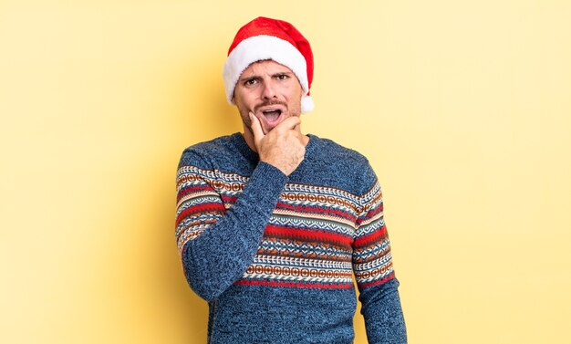 Junger gutaussehender Mann mit weit geöffnetem Mund und Augen und Hand am Kinn. Weihnachtskonzept