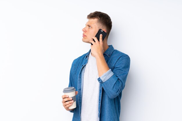 Junger gutaussehender Mann über dem Weiß, das Kaffee hält, um und ein Mobile wegzunehmen