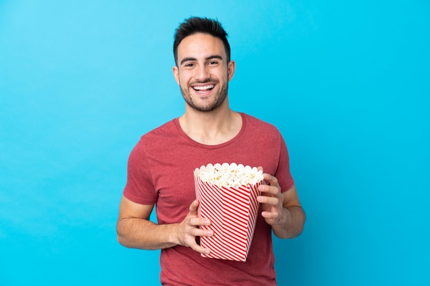 Junger gutaussehender Mann über der lokalisierten blauen Wand, die einen großen Eimer Popcorn hält