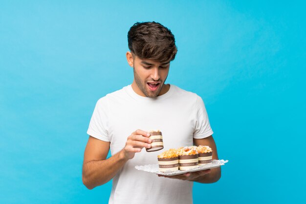 Junger gutaussehender Mann über der lokalisierten blauen Wand, die Minikuchen hält und überrascht