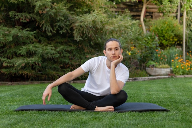 Junger gutaussehender Teenager-Mann, der in einem Yoga-Kurs in die Kamera schaut und seinen Kopf mit gekreuzten Beinen auf seine Hand legt