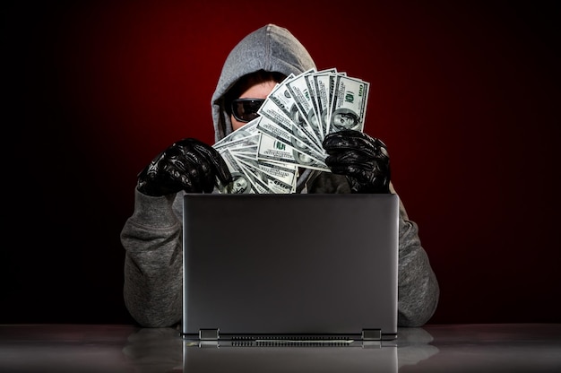 Junger Hacker in einer Sonnenbrille mit Laptop und Geld in der Hand