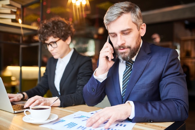 Junger Händler in Abendgarderobe berät einen der Kunden am Telefon, während er Finanzdiagramme im Café analysiert